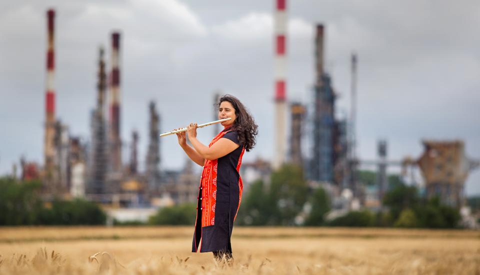 Portrait de Naïssam Jalal jouant de la flûte traversière dans un champ de blé, une usine en arrière plan