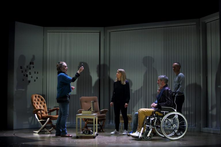 Des personnes dans une pièce. Une femme montre une photo, les autres, dont une personne en fauteuil roulant, regardent.